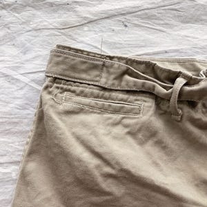 May include: Close-up of khaki-colored shorts, showcasing the waistband, belt loops, and a pocket. The fabric appears to be a sturdy cotton blend. The shorts have a tie closure and a classic design.
