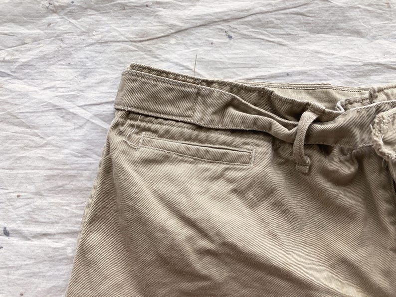 May include: Close-up of khaki-colored shorts, showcasing the waistband, belt loops, and a pocket. The fabric appears to be a sturdy cotton blend. The shorts have a tie closure and a classic design.