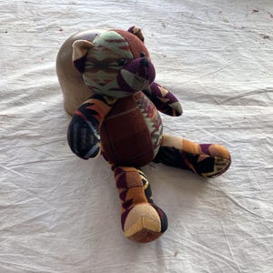 Può includere: Un orsacchiotto fatto a mano con un design patchwork. L'orso presenta vari tessuti fantasia nei toni del marrone, arancione, viola e verde. Ha un naso marrone scuro e occhi neri. L'orso è appoggiato su una superficie bianca.