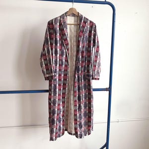 May include: A long, open-front robe with a plaid pattern in shades of red, white, and grey hangs on a wooden hanger. The robe is displayed on a blue metal clothing rack against a white wall. The robe has long sleeves and a relaxed fit.