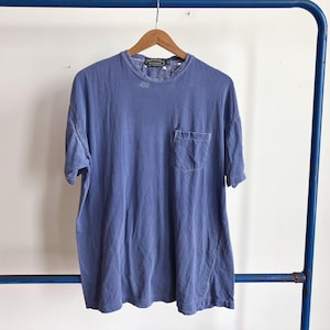 May include: A faded periwinkle blue t-shirt with a chest pocket hangs on a wooden hanger. The shirt has short sleeves and a crew neck. The garment is displayed against a white background and a blue metal rack.