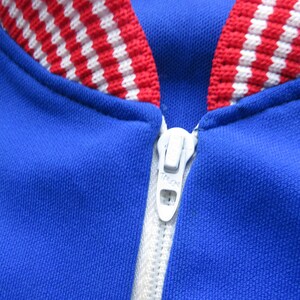 May include: A blue jacket with a red and white striped collar and a white zipper.