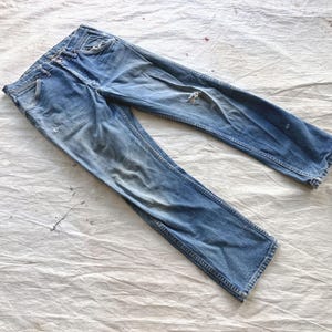 Peut inclure: Jean en denim bleu clair d&eacute;lav&eacute; avec des d&eacute;tails us&eacute;s. Le jean a une conception classique &agrave; cinq poches, une braguette boutonn&eacute;e et une coupe droite. Le tissu pr&eacute;sente des signes d'usure, avec une petite d&eacute;chirure sur la jambe droite.