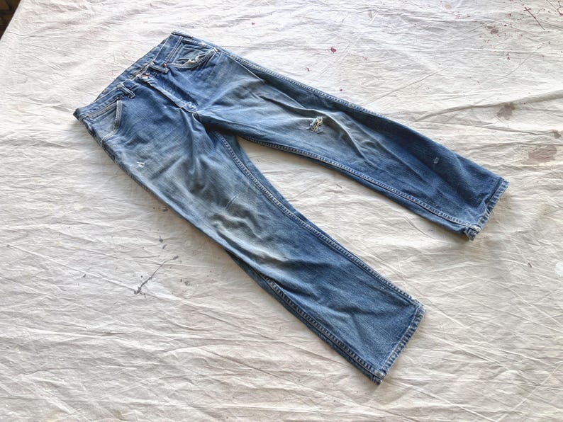 Peut inclure: Jean en denim bleu clair d&eacute;lav&eacute; avec des d&eacute;tails us&eacute;s. Le jean a une conception classique &agrave; cinq poches, une braguette boutonn&eacute;e et une coupe droite. Le tissu pr&eacute;sente des signes d'usure, avec une petite d&eacute;chirure sur la jambe droite.