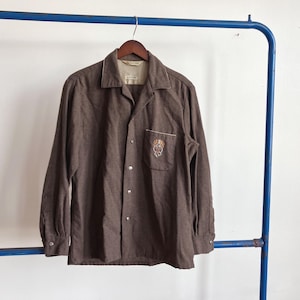 May include: Brown long-sleeve shirt with a camp collar and a chest pocket featuring an embroidered design. The shirt has a button-down front and cuffs. It is hanging on a wooden hanger.