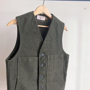 May include: A dark olive green wool vest featuring a V-neckline, a front button closure with four dark buttons, and two large patch pockets on the front. The garment is displayed on a wooden hanger and includes a small white label at the neck.