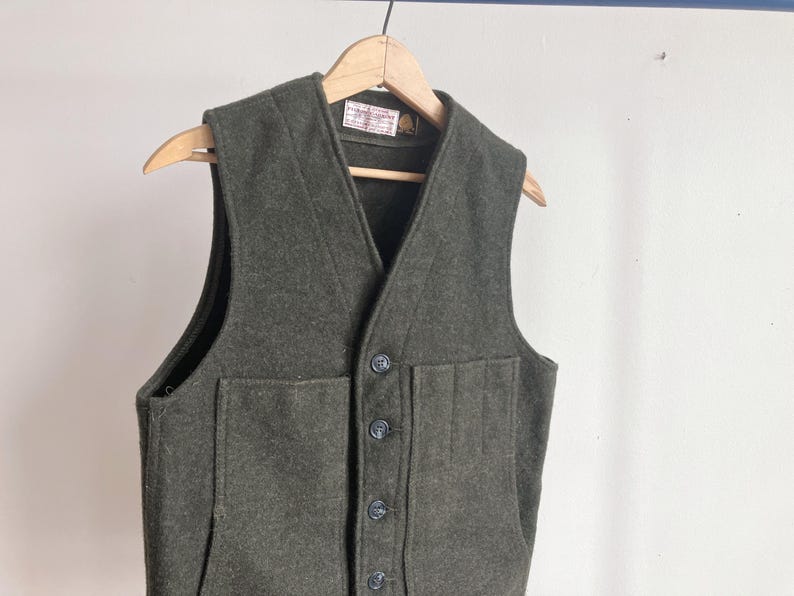 May include: A dark olive green wool vest featuring a V-neckline, a front button closure with four dark buttons, and two large patch pockets on the front. The garment is displayed on a wooden hanger and includes a small white label at the neck.