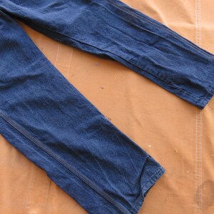 May include: A pair of dark blue denim jeans with a straight leg and a slightly faded appearance. The jeans are laid flat on a brown surface.