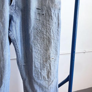 May include: Light blue denim jeans with distressed details, including faded areas and small tears. The jeans are hanging on a blue metal bar, showcasing the texture and wear of the fabric. The jeans appear to be vintage or have a worn-in look.