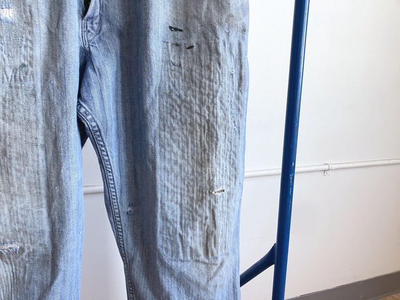 May include: Light blue denim jeans with distressed details, including faded areas and small tears. The jeans are hanging on a blue metal bar, showcasing the texture and wear of the fabric. The jeans appear to be vintage or have a worn-in look.