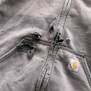 May include: A gray zip-up hoodie with a large, distressed hole in the fabric. The hoodie has a Carhartt logo patch on the front.
