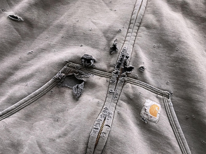 May include: A gray zip-up hoodie with a large, distressed hole in the fabric. The hoodie has a Carhartt logo patch on the front.