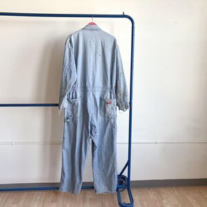 May include: Light blue denim jumpsuit hanging on a blue metal rack. The jumpsuit has long sleeves, multiple pockets, and a red logo patch. The garment is slightly distressed, with visible wear and tear.
