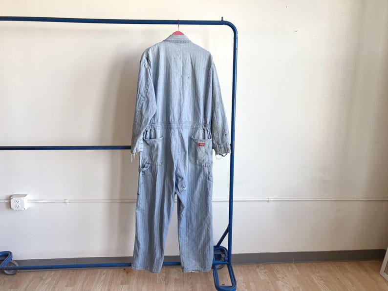May include: Light blue denim jumpsuit hanging on a blue metal rack. The jumpsuit has long sleeves, multiple pockets, and a red logo patch. The garment is slightly distressed, with visible wear and tear.