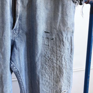 May include: Light blue denim jeans with a zipper fly. The jeans show signs of wear, with distressed areas and dark spots. The fabric has a textured appearance. The jeans are hanging on a blue metal bar.