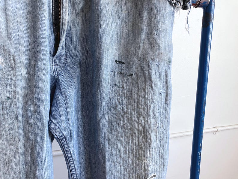 May include: Light blue denim jeans with a zipper fly. The jeans show signs of wear, with distressed areas and dark spots. The fabric has a textured appearance. The jeans are hanging on a blue metal bar.