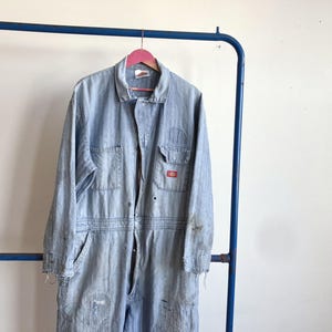May include: Light blue denim work jumpsuit with multiple pockets and a zipper closure. The jumpsuit is hanging on a pink hanger. The Dickies logo is visible on the chest pocket. The garment shows signs of wear and tear.