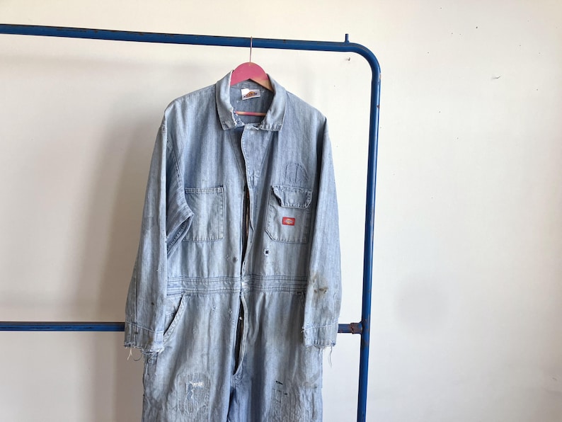 May include: Light blue denim work jumpsuit with multiple pockets and a zipper closure. The jumpsuit is hanging on a pink hanger. The Dickies logo is visible on the chest pocket. The garment shows signs of wear and tear.