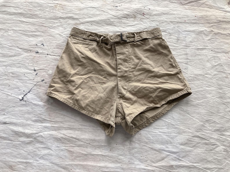 May include: Tan-colored shorts with a belt and buckle closure. The shorts have a high waist and a classic design. The fabric appears to be a sturdy cotton or blend. The shorts are laid flat on a textured, off-white surface.