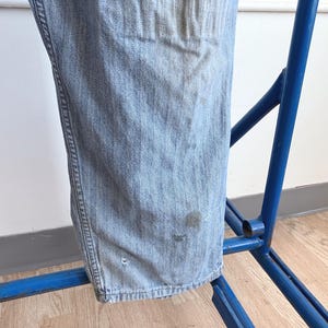 May include: Light blue denim jeans hanging on a blue metal rack. The jeans show signs of wear with some discoloration and small imperfections. The fabric has a classic denim texture.