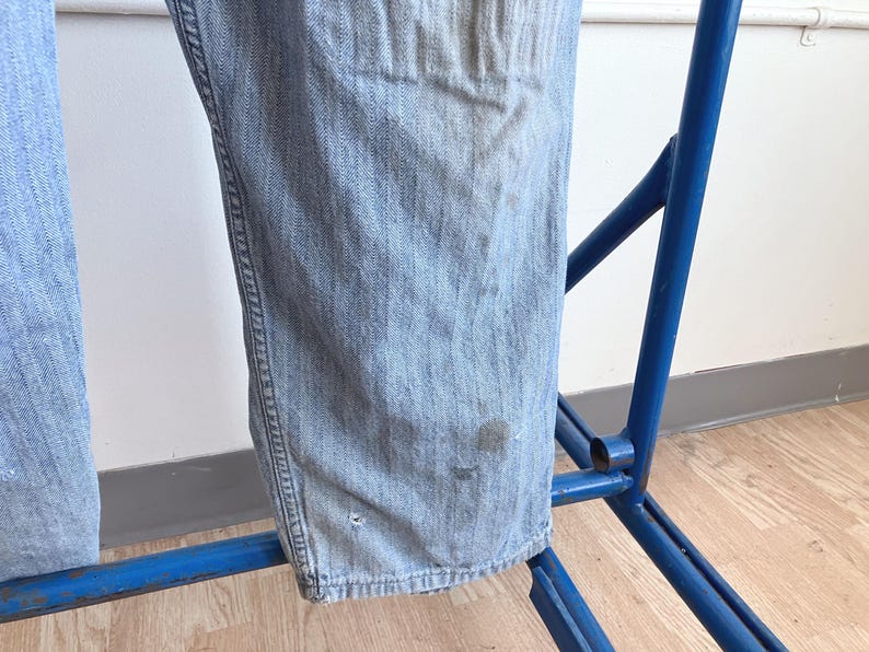 May include: Light blue denim jeans hanging on a blue metal rack. The jeans show signs of wear with some discoloration and small imperfections. The fabric has a classic denim texture.