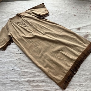 Puede incluir: Un vestido color beige, de manga corta, con cuello en V y flecos. El vestido tiene cierre de botones y detalles de flecos en las mangas y el dobladillo. La tela parece ser un material ligero, adecuado para climas cálidos.