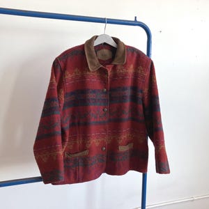 Puede incluir: Chaqueta estampada en rojo y azul con cuello y detalles de bolsillo de ante marrón. La chaqueta está abotonada y colgada de una percha blanca. La chaqueta se exhibe en una percha de metal azul.