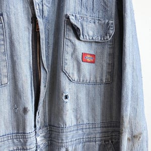 May include: Light blue denim work jumpsuit with a front zipper and multiple pockets. Features a small red and white Dickies logo patch on the chest pocket. The jumpsuit shows signs of wear and tear, with some discoloration and small holes.