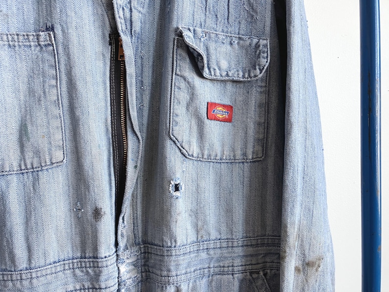 May include: Light blue denim work jumpsuit with a front zipper and multiple pockets. Features a small red and white Dickies logo patch on the chest pocket. The jumpsuit shows signs of wear and tear, with some discoloration and small holes.