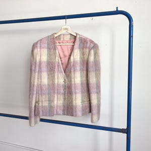 Puede incluir: Blazer vintage a cuadros en tonos pastel, con un aspecto suave y texturizado. La chaqueta presenta un diseño de cuello en V, dos bolsillos delanteros y un cierre de un solo botón. Cuelga de una percha de madera suspendida de un perchero de metal azul.
