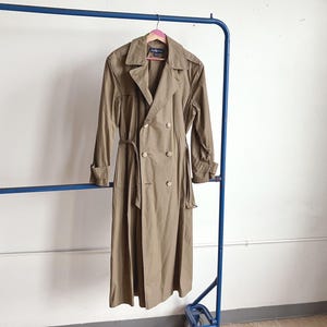 May include: A double-breasted trench coat in a muted olive-brown tone, featuring a wide collar, long sleeves with adjustable cuffs, and a matching waist belt. The coat has a button-front closure and a structured silhouette, hanging on a hanger.