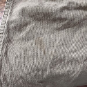 May include: Close-up of tan fabric with visible diagonal weave and a brown stain. The edge of the fabric shows a seam with double stitching. The fabric appears to be a durable material, possibly for pants or a similar garment.