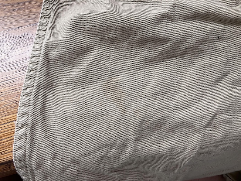 May include: Close-up of tan fabric with visible diagonal weave and a brown stain. The edge of the fabric shows a seam with double stitching. The fabric appears to be a durable material, possibly for pants or a similar garment.