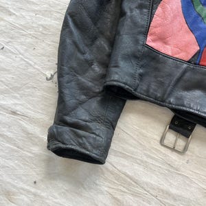 Op de afbeelding: Zwarte leren jas met een kleurrijk abstract ontwerp op de bovenrug. De jas heeft een gespriem en een klassieke kraag. Het leer lijkt versleten, wat het een vintage uitstraling geeft. De mouw is zichtbaar en toont de manchet.