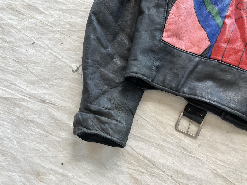 Op de afbeelding: Zwarte leren jas met een kleurrijk abstract ontwerp op de bovenrug. De jas heeft een gespriem en een klassieke kraag. Het leer lijkt versleten, wat het een vintage uitstraling geeft. De mouw is zichtbaar en toont de manchet.
