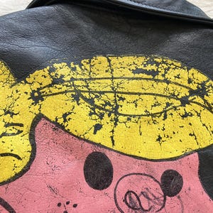 Op de afbeelding: Close-up van een zwarte leren jas met een distressed cartoon varkensontwerp. Het varken heeft een roze huid, geel haar en zwarte accenten. Het leer van de jas vertoont slijtage.