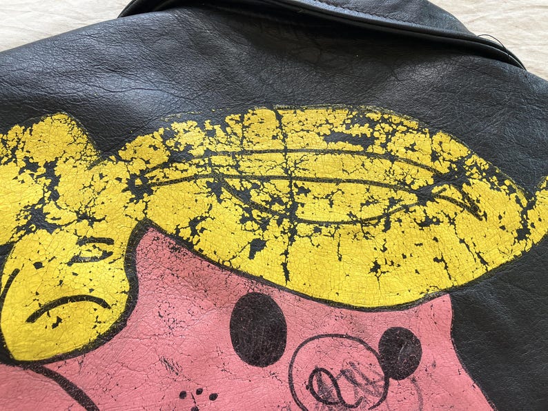 Op de afbeelding: Close-up van een zwarte leren jas met een distressed cartoon varkensontwerp. Het varken heeft een roze huid, geel haar en zwarte accenten. Het leer van de jas vertoont slijtage.