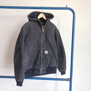 May include: A charcoal gray hooded jacket with a front zipper closure, long sleeves, and ribbed cuffs. The jacket features a front pouch pocket with a small rectangular Carhartt brand patch sewn onto the front. The hood includes a drawstring.
