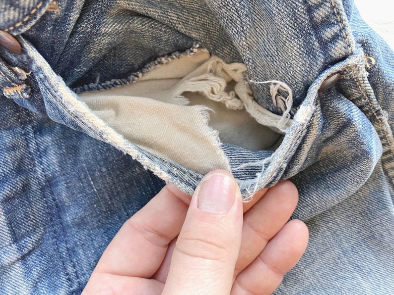 Peut inclure: Gros plan sur une poche de jean en denim vieilli. La doublure de la poche est visible, montrant des bords effiloch&eacute;s et un aspect us&eacute;. Le denim est un d&eacute;lavage bleu clair, avec une usure visible.
