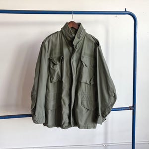May include: Olive green military-style jacket with multiple front pockets and a high collar. The jacket is hanging on a wooden hanger suspended from a blue metal clothing rack. The jacket is made of a durable fabric.