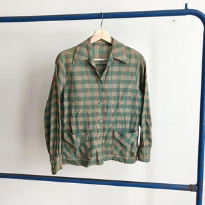 May include: A vintage button-down shirt in a green and tan plaid pattern. The long-sleeved shirt has two front pockets and a collared neckline. The shirt is hanging on a wooden hanger on a blue metal clothing rack.