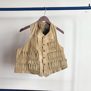 Puede incluir: Un chaleco de color beige con múltiples bolsillos, colgado de una percha de madera. El chaleco tiene una parte delantera abotonada y está hecho de un material texturizado. Se muestra sobre un fondo blanco.