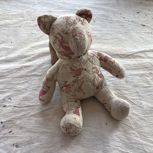 Pode incluir: Um urso de pelúcia feito à mão com um design acolchoado. O urso é bege com um padrão de figuras rosa e vermelhas. Possui um rosto costurado e está sobre uma superfície branca.