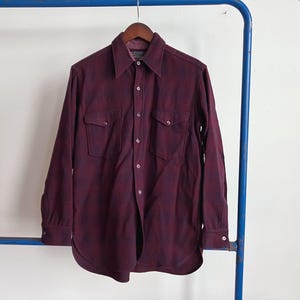 May include: A burgundy and navy plaid button-down shirt with two chest pockets. The long-sleeved shirt is hanging on a wooden hanger against a white background. The shirt has a classic collar and buttoned cuffs.