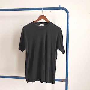 Könnte beinhalten: Ein schwarzes geripptes Kurzarm-Shirt, das an einem Holzbügel hängt. Das Shirt wird vor einer weißen Wand und einem blauen Metallgestell präsentiert. Das Shirt hat einen Rundhalsausschnitt und ein Etikett auf dem Rücken.