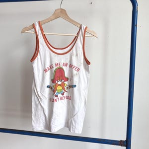 May include: A white sleeveless tank top with orange trim around the neckline and armholes. The front features a graphic of the cartoon character Yosemite Sam holding two pistols, with the text "MAKE ME AN OFFER I CAN'T REFUSE" printed in red.
