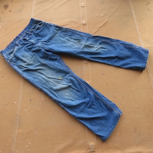 May include: A pair of blue denim jeans with a faded look and some wear and tear. The jeans are laid out flat on a brown surface.