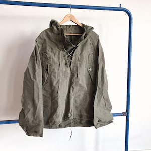 Könnte beinhalten: Eine olivgrüne Kapuzenjacke zum Überziehen mit Schnürung an der Vorderseite, passenden Stoffbändern, langen Ärmeln mit geknöpften Manschetten und zwei Vordertaschen mit geknöpften Klappen. Die Jacke ist aus einem strapazierfähigen Baumwollstoff.