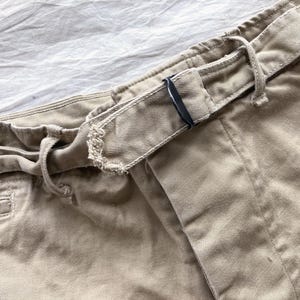 May include: Close-up of khaki-colored pants with a drawstring waist and a belt loop. The belt loop has a black metal buckle. The fabric appears to be a sturdy cotton or blend, suitable for casual wear.