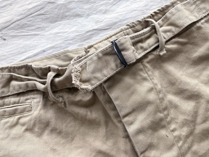 May include: Close-up of khaki-colored pants with a drawstring waist and a belt loop. The belt loop has a black metal buckle. The fabric appears to be a sturdy cotton or blend, suitable for casual wear.
